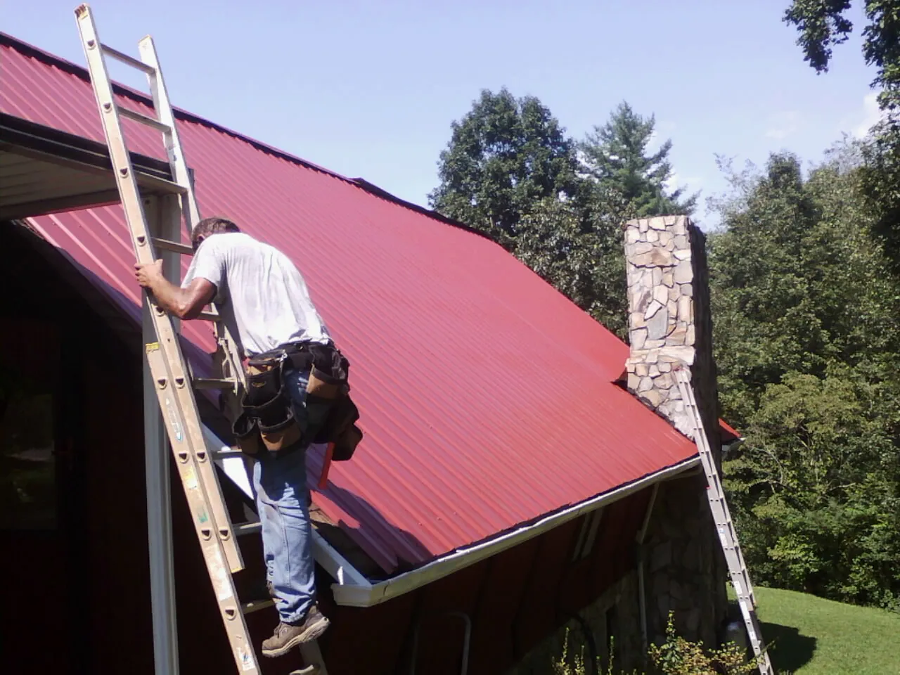 Roofer on a ladder beside a red metal roof and stone chimney — Soffit & Fascia Repair by Roofing Brothers in Bloomington